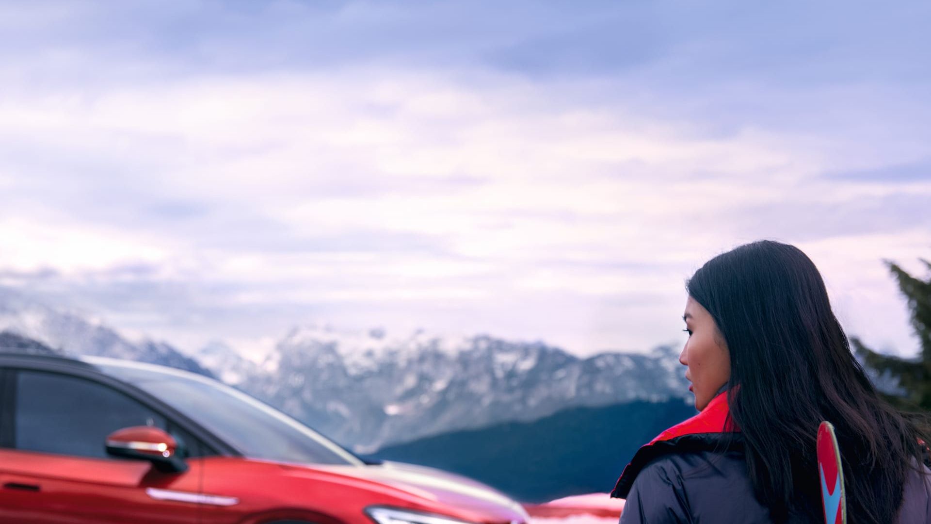 Vista lateral de um VW ID.4 GTX vermelho perante uma montanha com uma mulher a olhar para o carro em primeiro plano