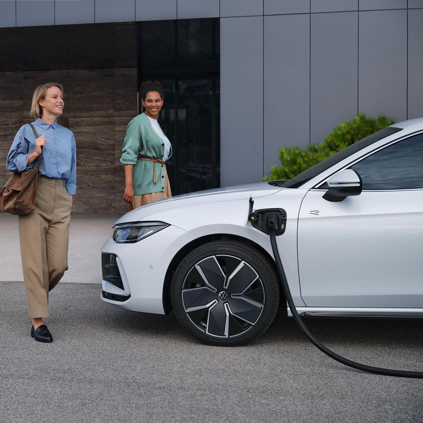 VW Passat branco lateralmente a um edifício de escritórios com o cabo de carregamento ligado, duas mulheres aproximam-se do carro.