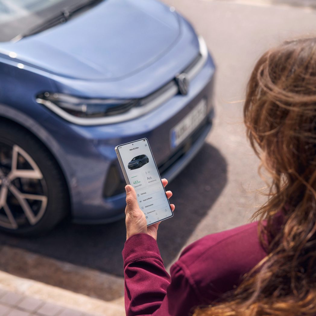 Eine Frau schaut auf ihr Smartphone, vor ihr parkt ein blauer VW ID.3 Neo