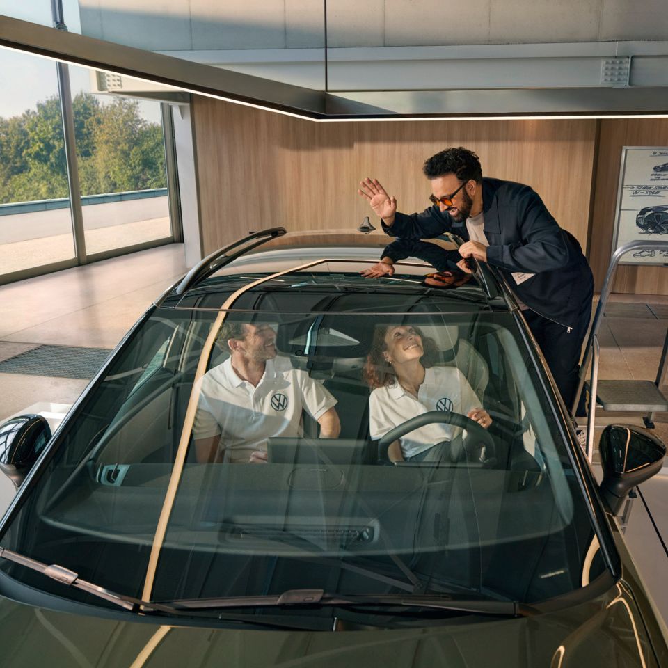Um homem encontra-se numa escada ao lado de um VW ID. Cross a acenar pelo teto panorâmico às pessoas sentadas no seu interior.
