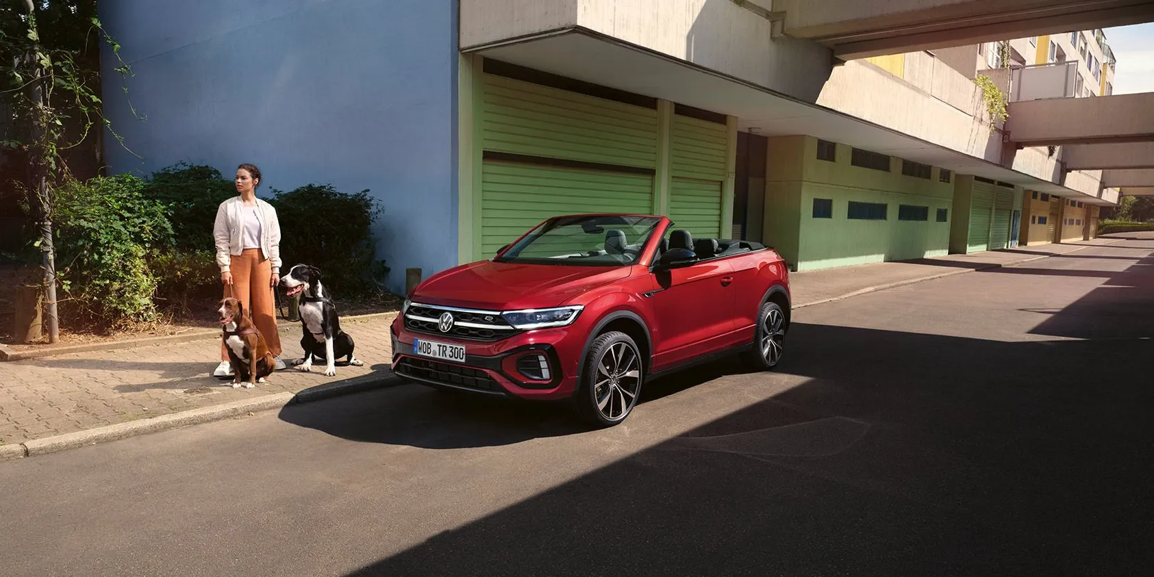 VW T-Roc Cabrio vermelho com a capota aberta, estacionado à beira da estrada. Uma mulher com dois cães está ao lado.