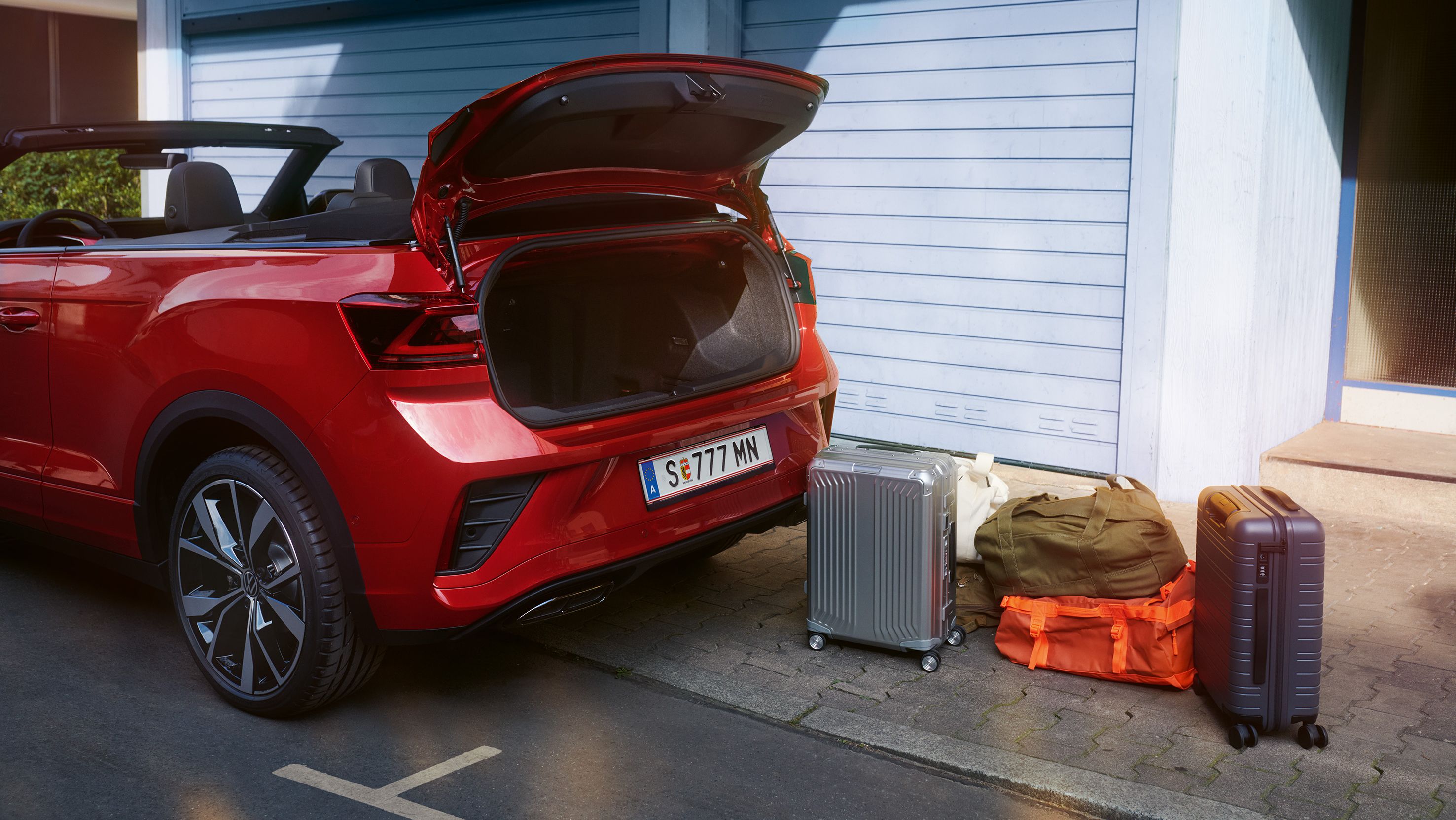 Ein VW T-Roc Cabriolet in rot parkt am Straßenrand. Blick seitlich auf den geöffneten Kofferraum, es stehen zwei Koffer und zwei Reisetaschen daneben. 