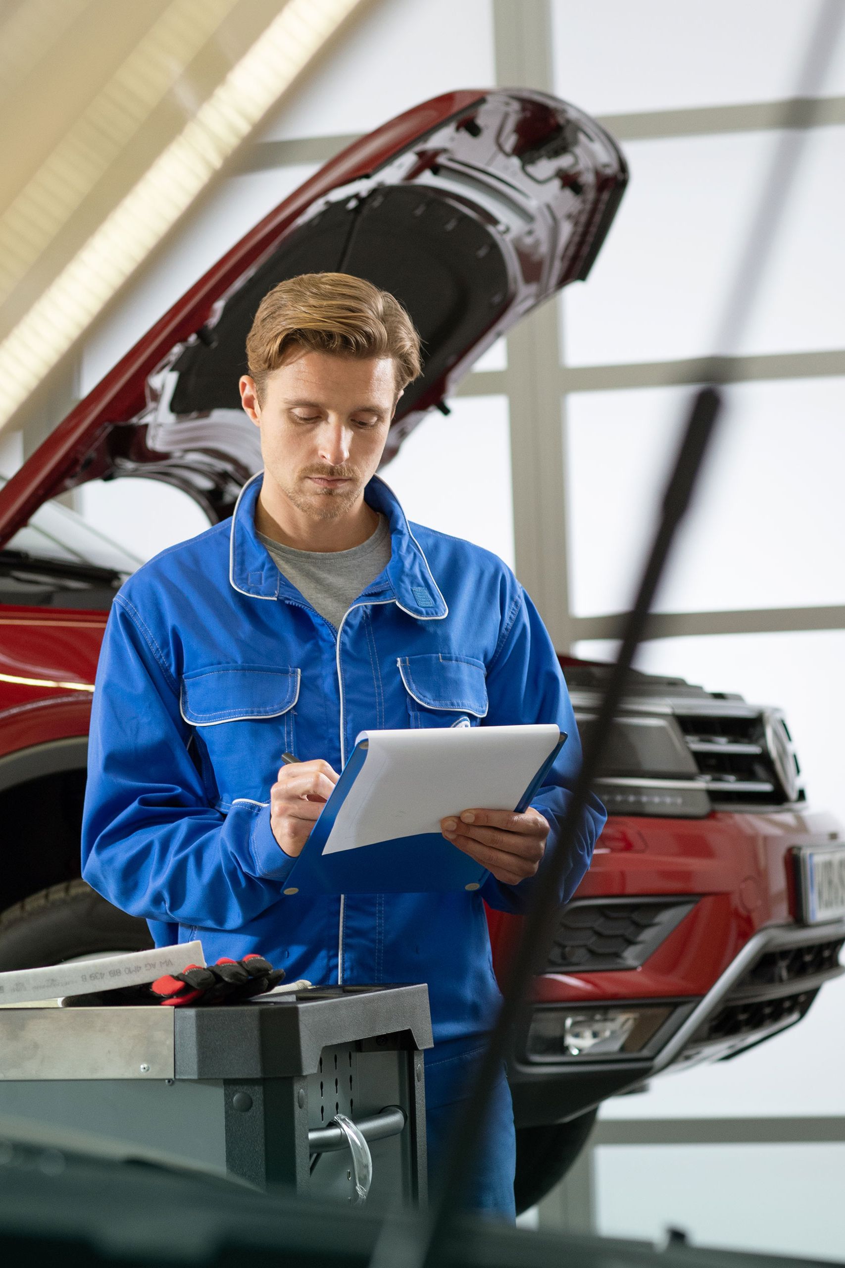 Funcionário do serviço VW verifica uma lista de verificação em frente de um VW vermelho numa rampa hidráulica com capô aberto