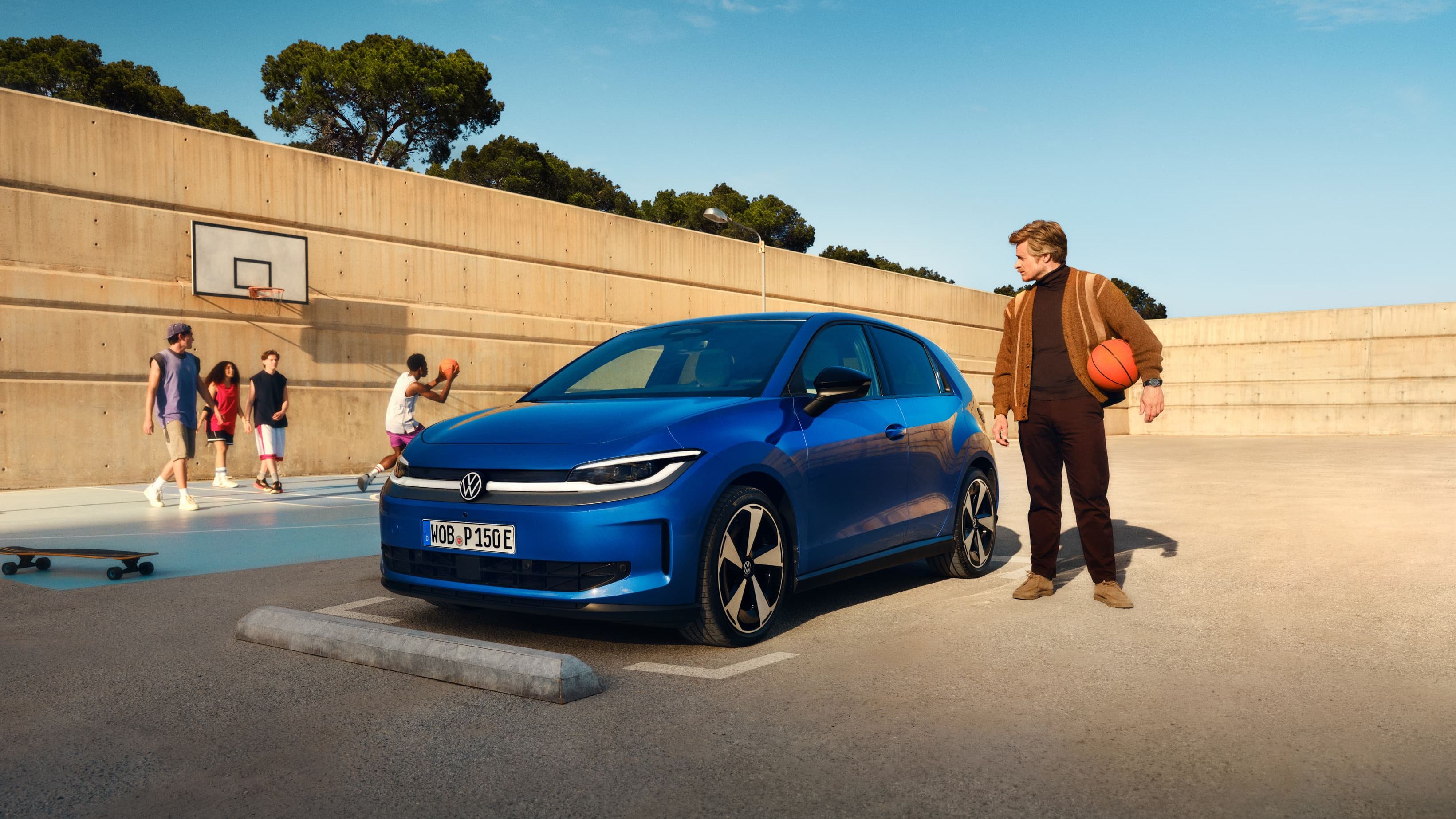 Um VW ID. Polo azul estacionado junto a um campo de basquete. Um homem está de pé nas proximidades, segura uma bola de basquetebol debaixo do braço.