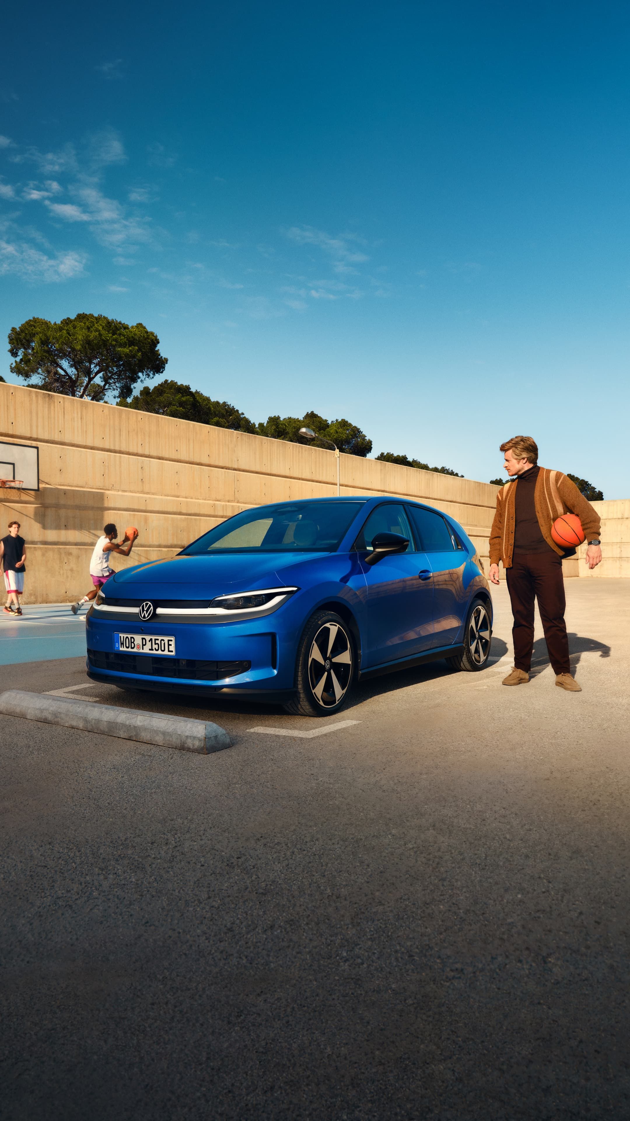 Um VW ID. Polo azul estacionado junto a um campo de basquete. Um homem está de pé nas proximidades, segura uma bola de basquetebol debaixo do braço.