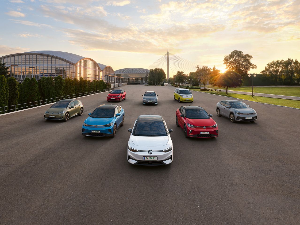 Fila de oito carros VW elétricos modernos e coloridos estacionados em formação simétrica numa ampla área asfaltada ao ar livre. Ao fundo há prédios de arquitetura curva com fachadas envidraçadas, cercados por árvores e um grande gramado. O céu ao entardecer exibe nuvens suaves iluminadas por tons quentes de laranja e amarelo.