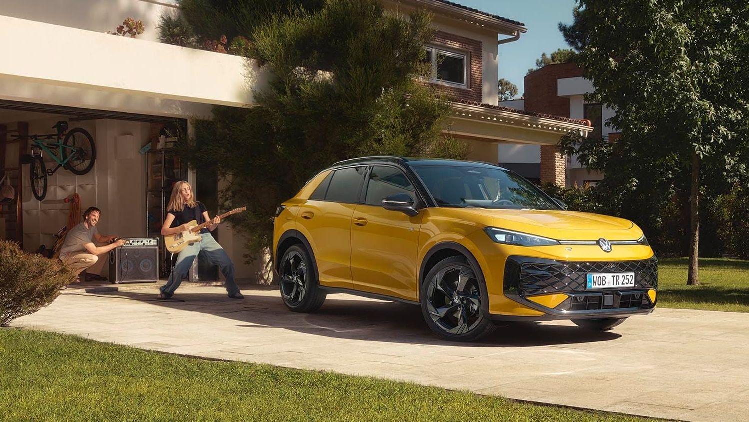 Um VW T-Roc R-Line amarelo em frente à garagem, uma jovem a tocar guitarra com o pai a controlar o altifalante.