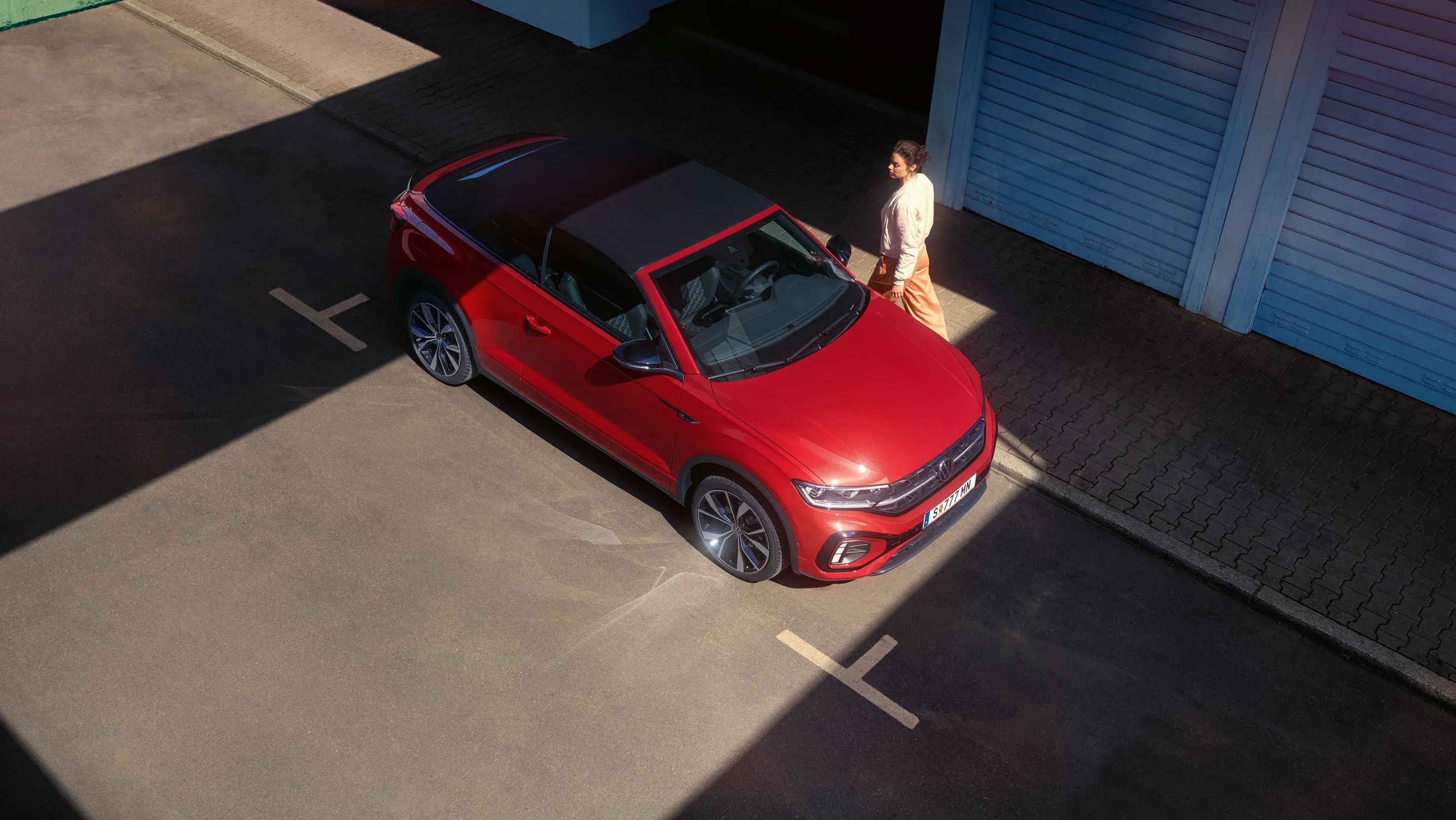 Vista aérea do VW T-Roc Cabrio vermelho com o tejadilho aberto. Estacionado à beira da estrada, uma mulher passa ao lado. 