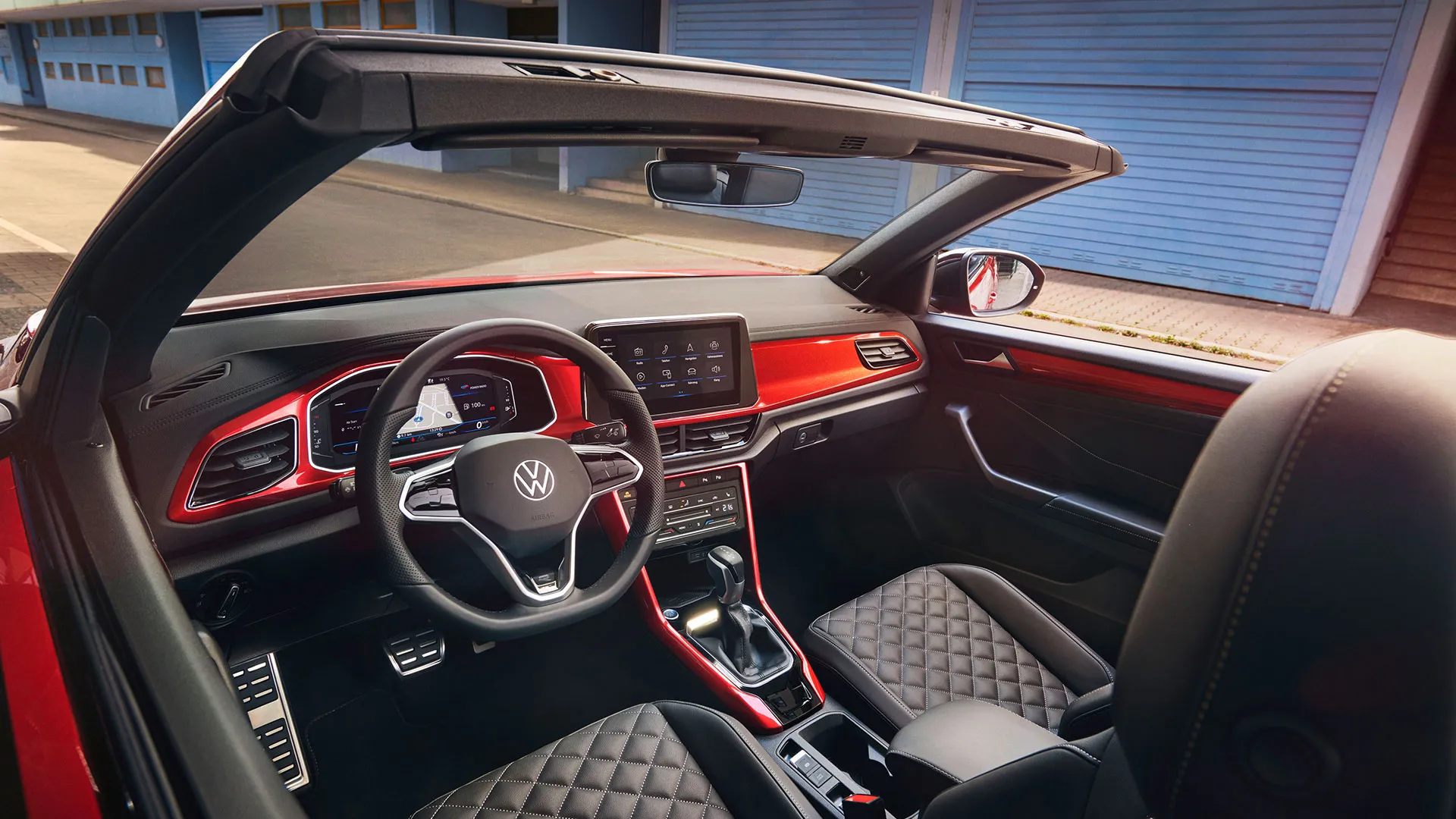 Interieur des VW T-Roc Cabriolet: Blick durch das geöffnete Dach von oben auf das Cockpit mit rotem Dekor und die vorderen Sitze. 