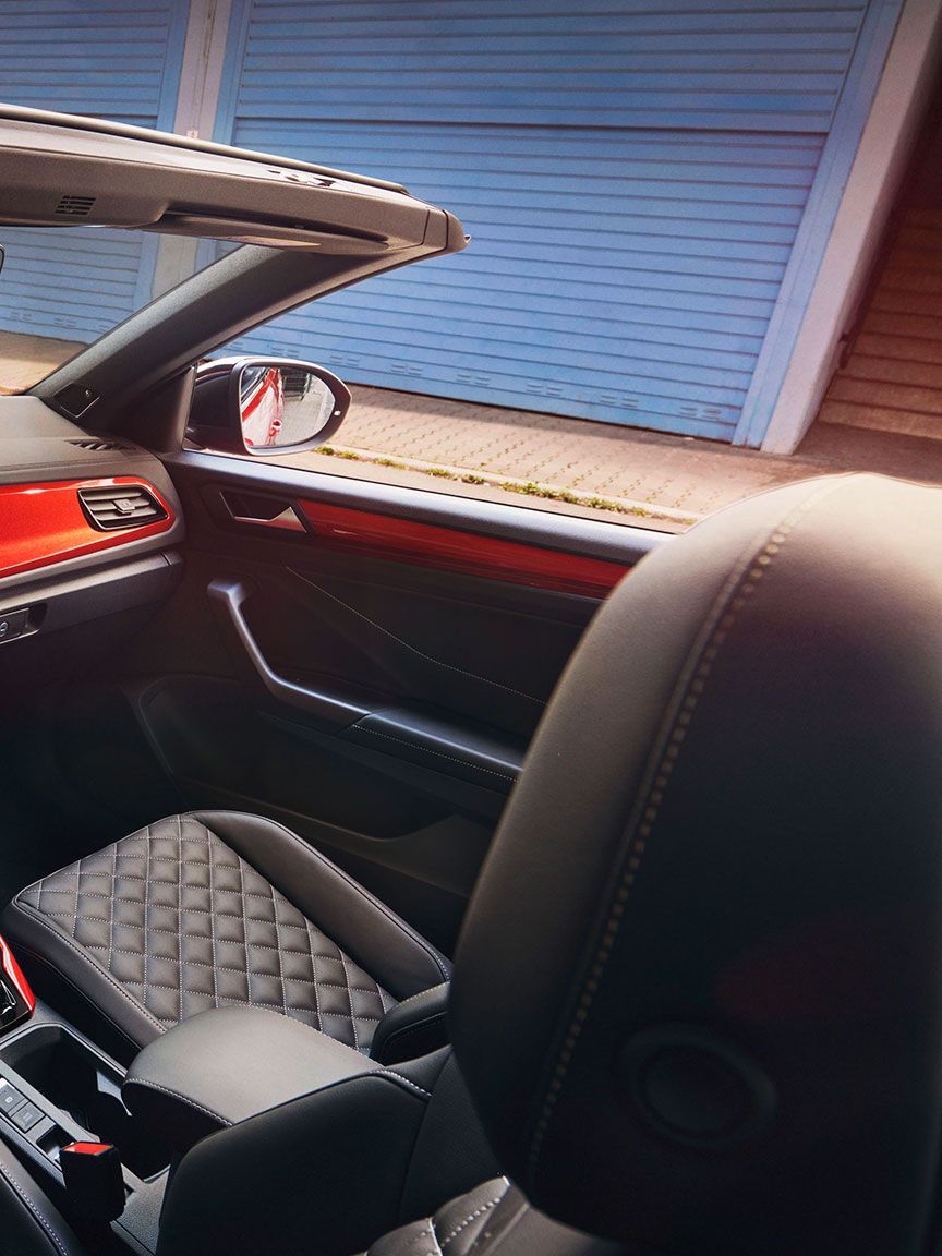 Interieur des VW T-Roc Cabriolet: Blick durch das geöffnete Dach von oben auf das Cockpit mit rotem Dekor und die vorderen Sitze. 