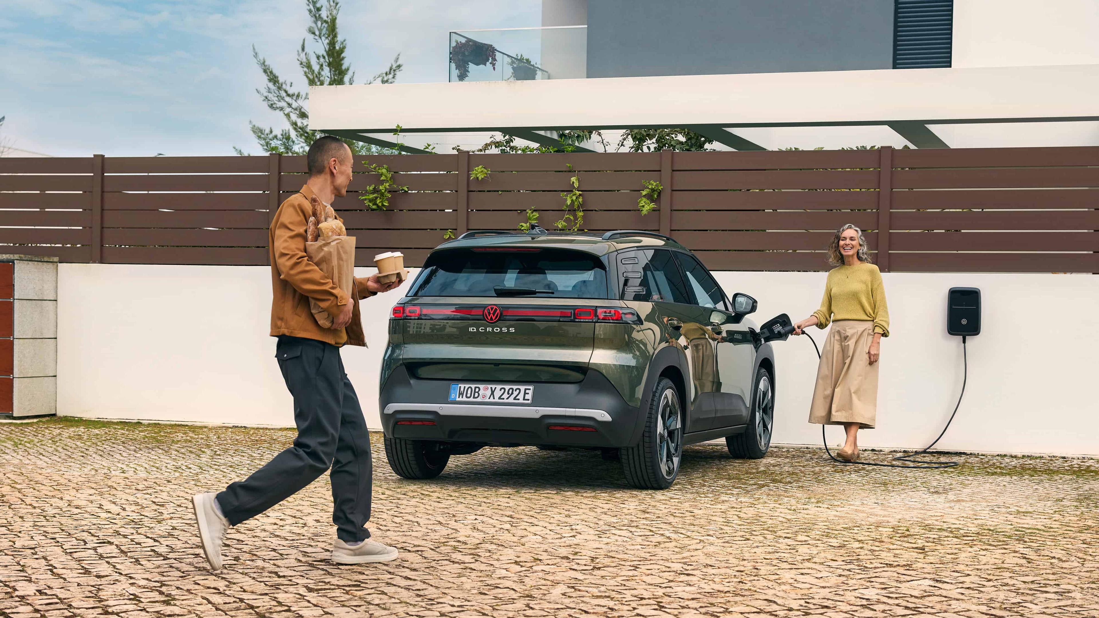 Homem caminha em direção a uma casa moderna enquanto segura um copo de café e pão. Mulher a inserir o carregador da wallbox de casa no VW ID. Cross   