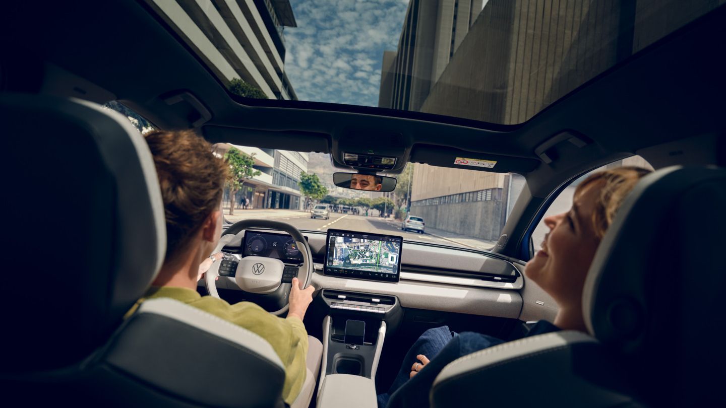 Um homem conduz um Volkswagen ID. Polo, no banco do passageiro uma mulher a rir. Veem-se edifícios altos através do teto de vidro panorâmico.