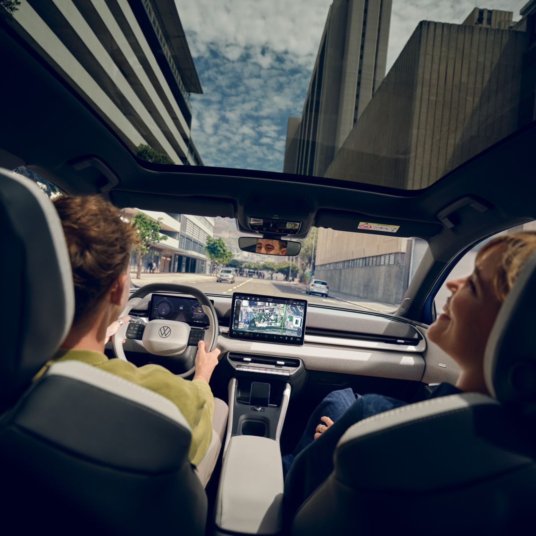 Um homem conduz um Volkswagen ID. Polo, no banco do passageiro uma mulher a rir. Veem-se edifícios altos através do teto de vidro panorâmico.