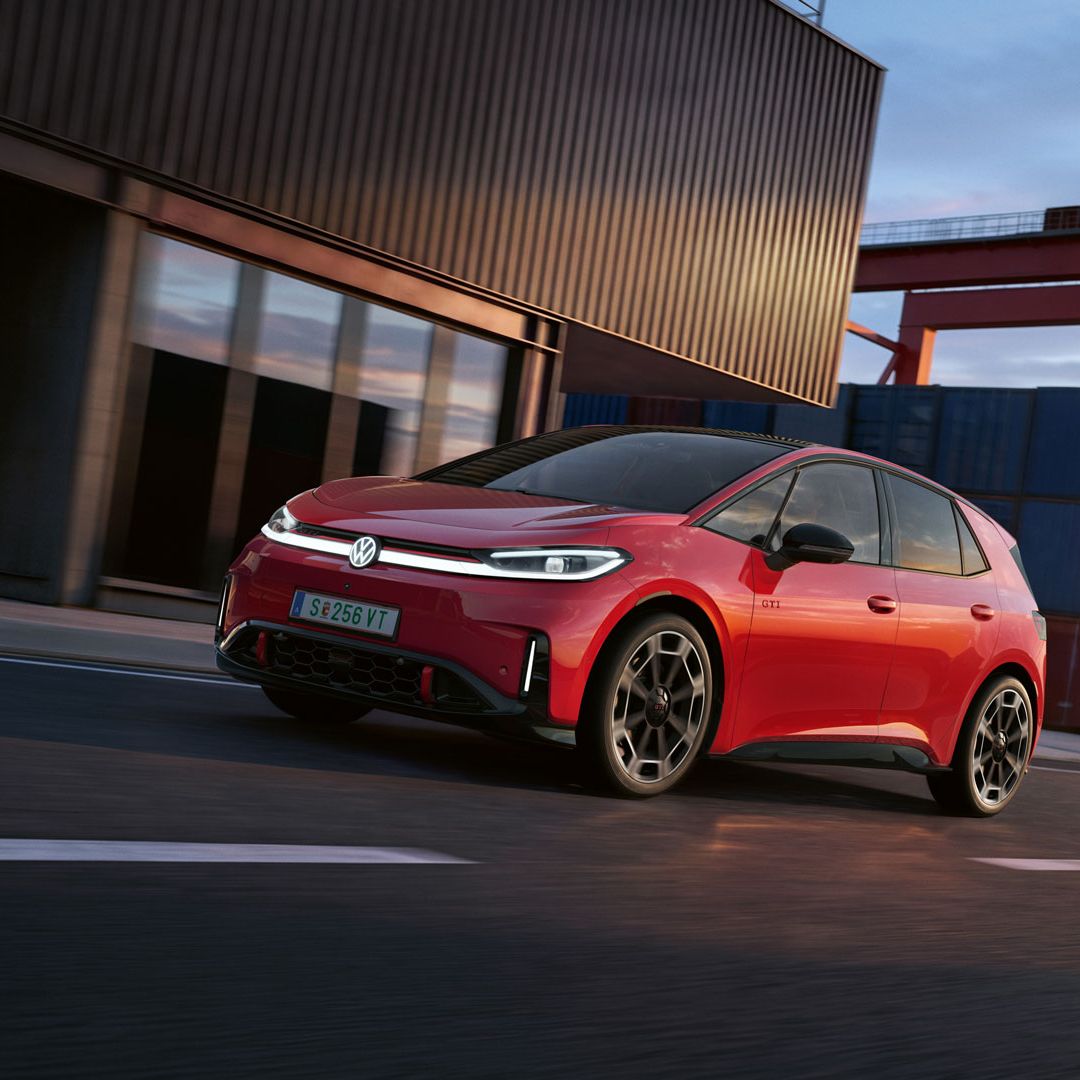 [AI-modified] – Um VW ID.3 GTI vermelho passa por um edifício portuário com os faróis ligados e o logótipo iluminado.