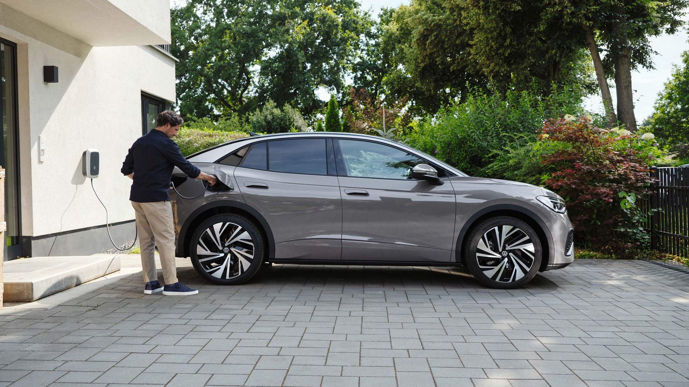 Homem parado ao lado do Volkswagen ID.5 em cor cinza, estacionado numa entrada de garagem pavimentada, carregando-o.