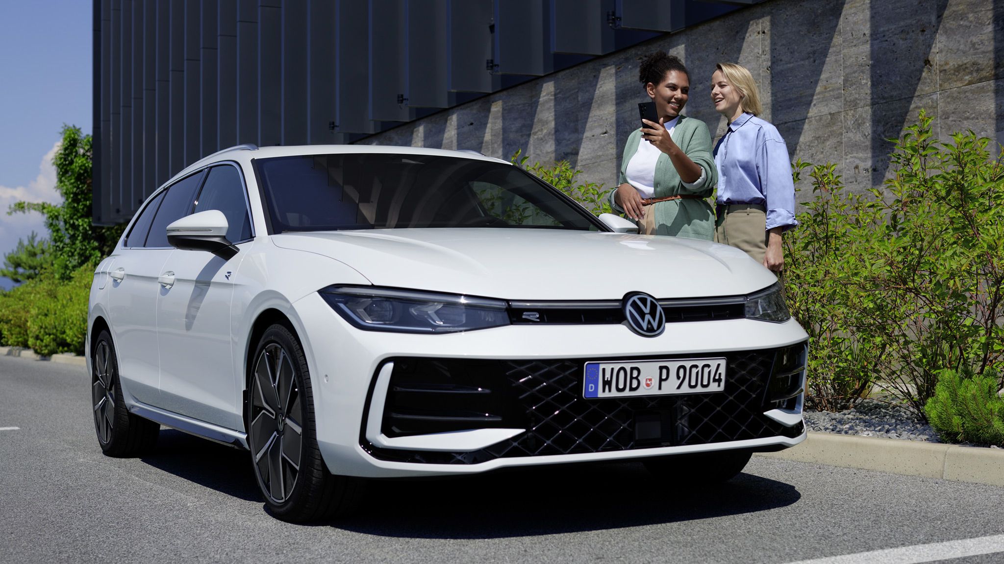 VW Passat branco estacionado lateralmente a um edifício, duas mulheres de pé do lado do condutor atrás dele, uma delas com o seu smartphone na mão.