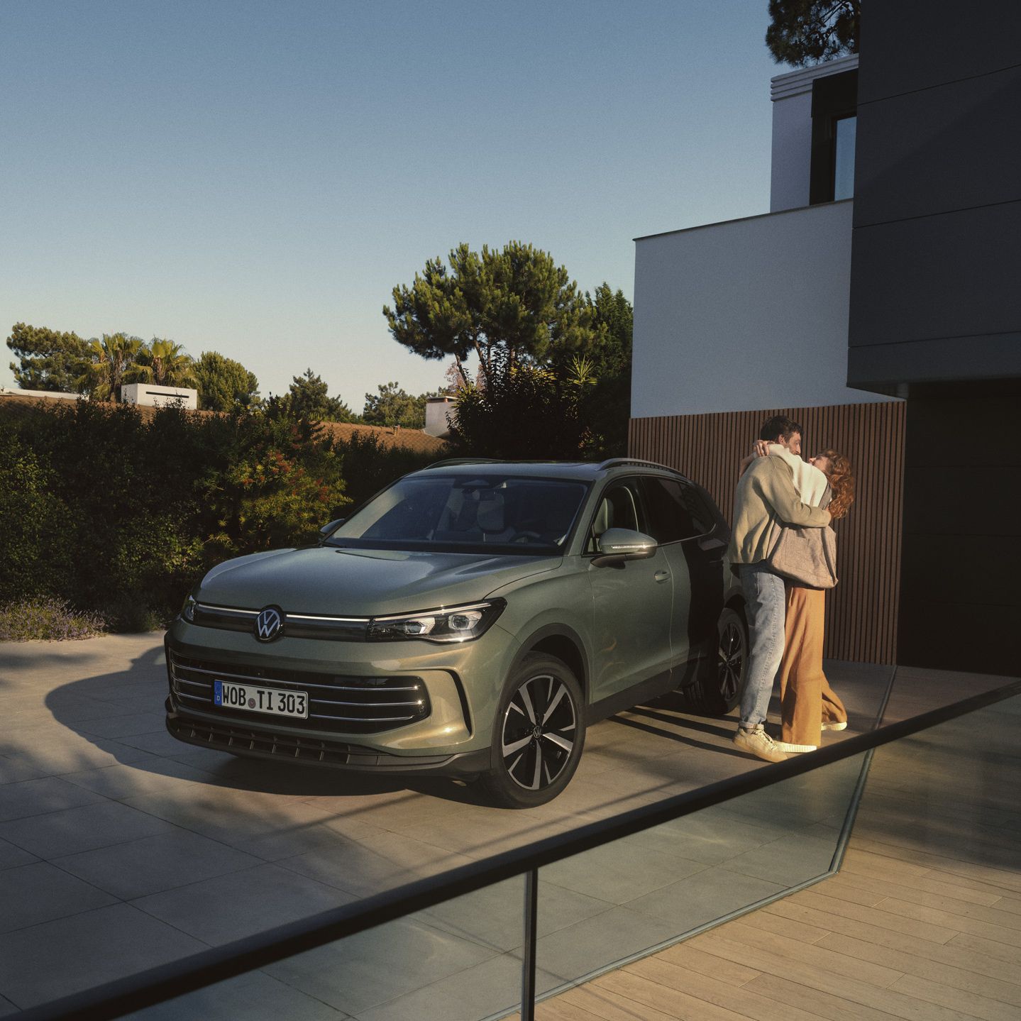 Vista frontal de um VW Tiguan verde estacionado na entrada de um edifício residencial moderno. Um jovem casal está ao lado dele e abraça-se.