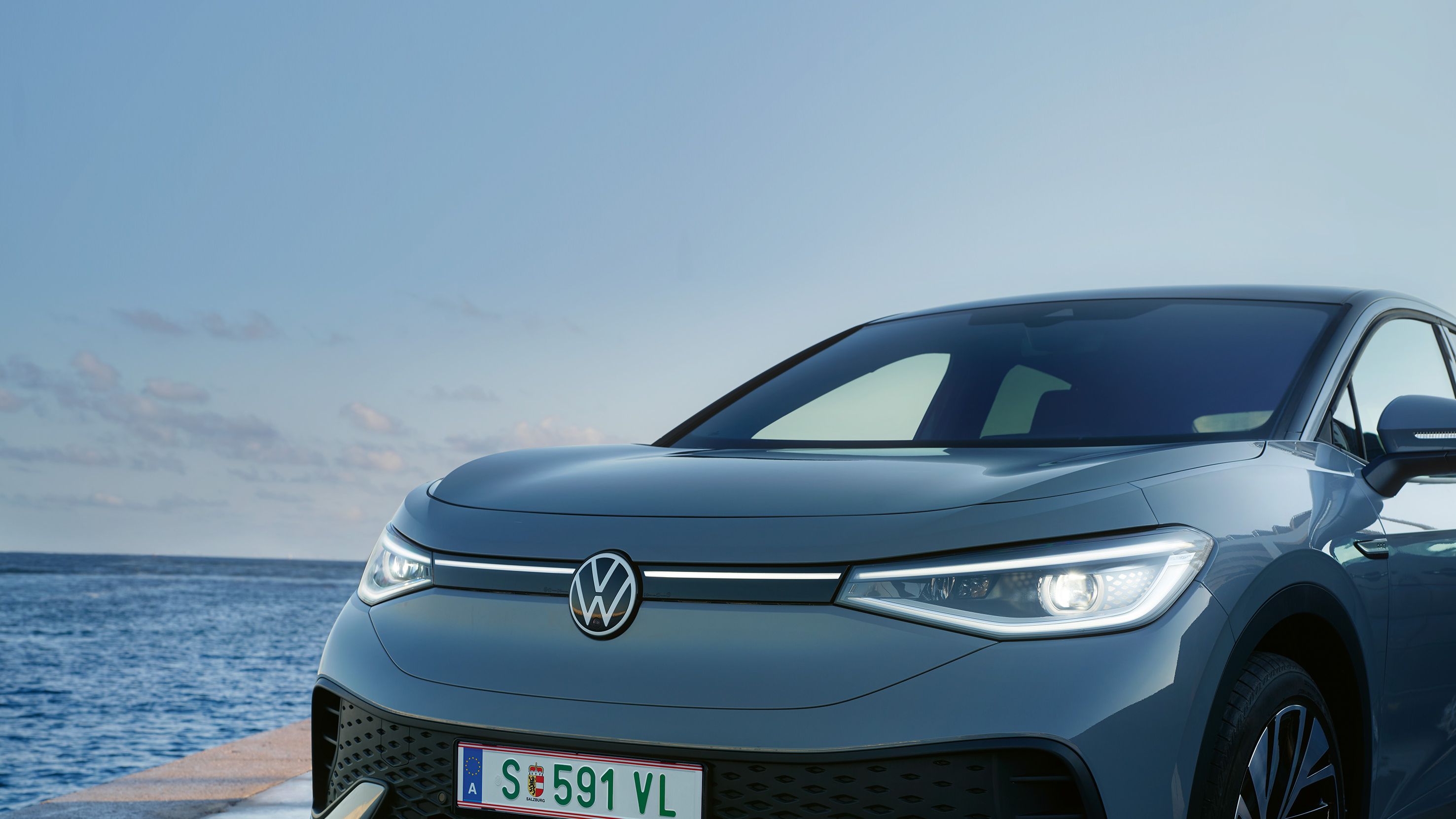 Dianteira de um VW ID.5 parado no cais de um porto ao entardecer, faróis LED acesos