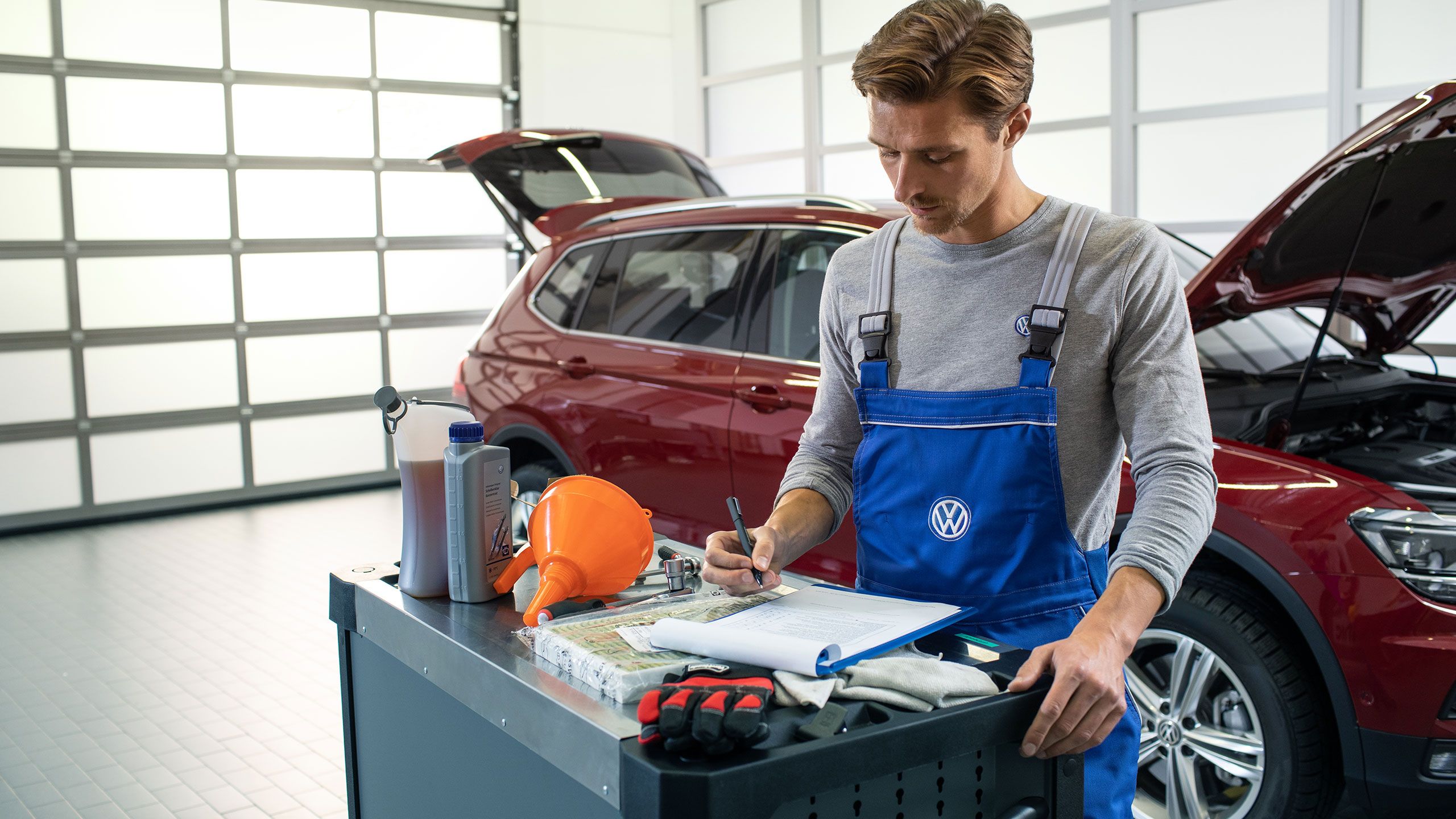 Um empregado do serviço VW está a consultar uma lista de verificação em frente de um VW Tiguan Allspace vermelho com capota aberta e compartimento de bagagem
