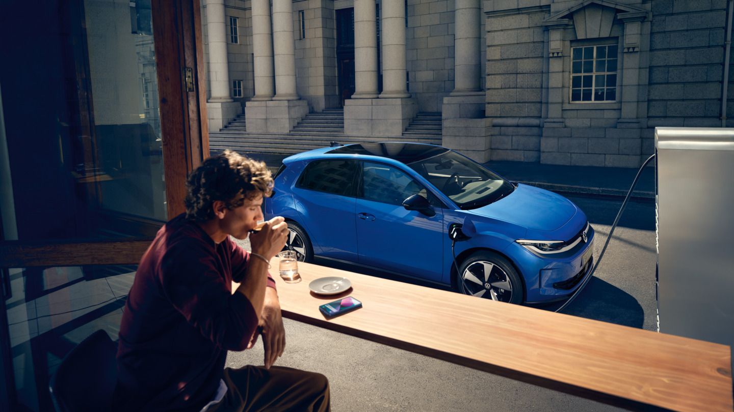 Um homem está sentado num café, através de uma janela aberta vê-se o VW ID. Polo a carregar numa estação de carregamento.