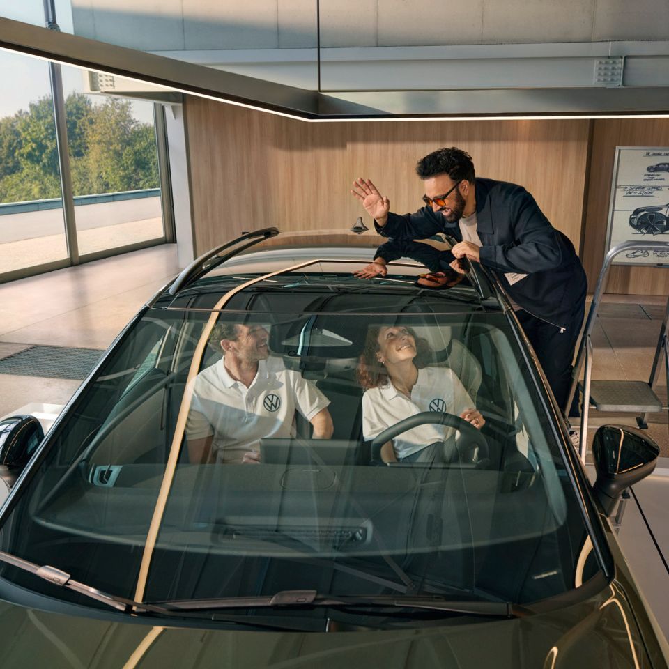 Um homem encontra-se numa escada ao lado de um VW ID. Cross a acenar pelo teto panorâmico às pessoas sentadas no seu interior.
