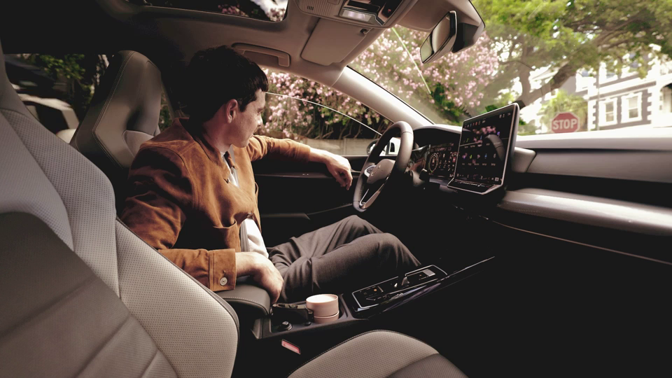 Ein Fahrer sitzt in einem modernen Auto vor einem Stoppschild. Die Person trägt eine braune Jacke und blickt nach hinten. Das Armaturenbrett zeigt ein großes Display.
