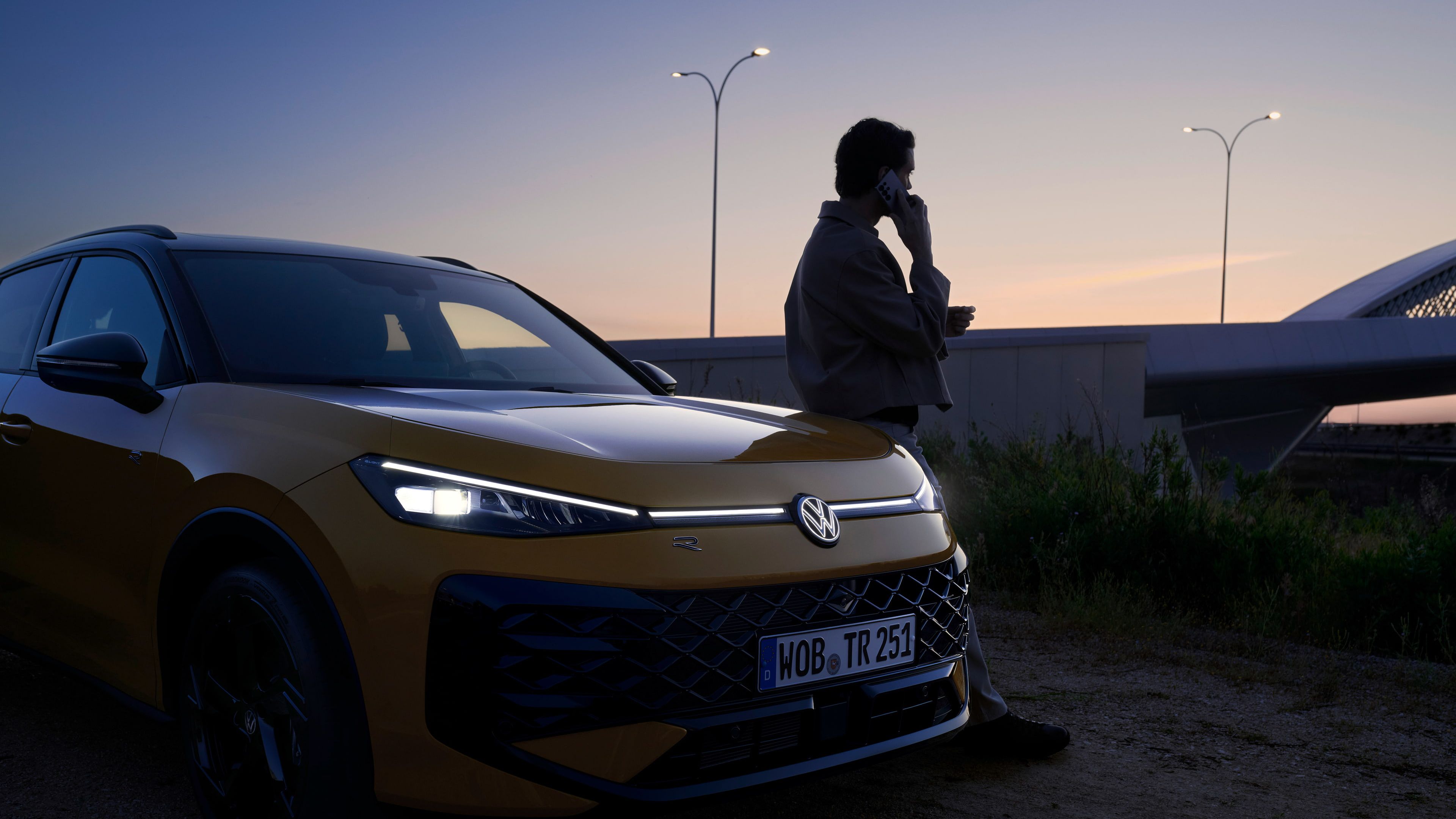 Um homem junto a um VW T-Roc R-Line com os faróis ligados e o logótipo iluminado.