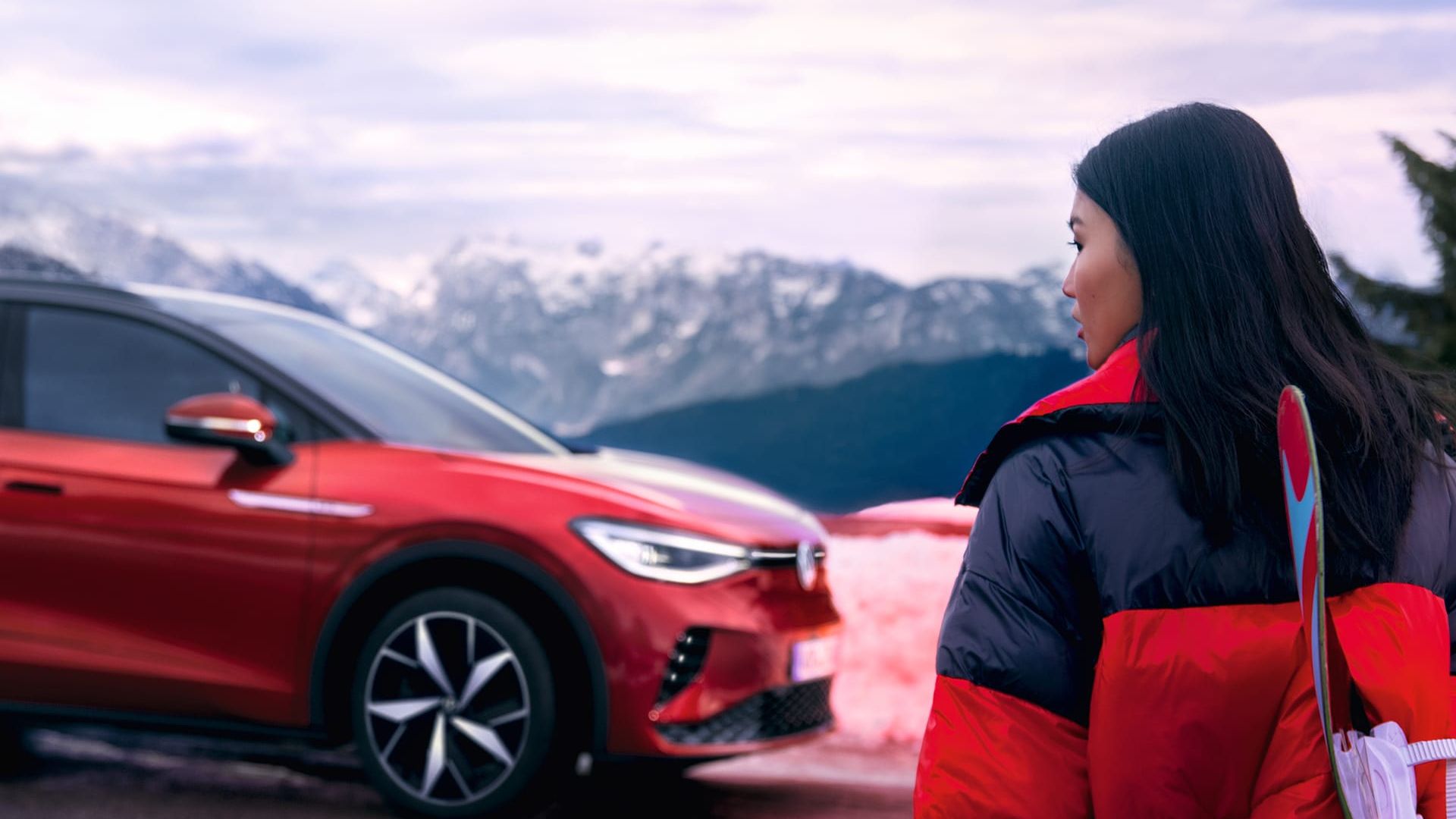 Vista lateral de um VW ID.4 GTX vermelho perante uma montanha com uma mulher a olhar para o carro em primeiro plano