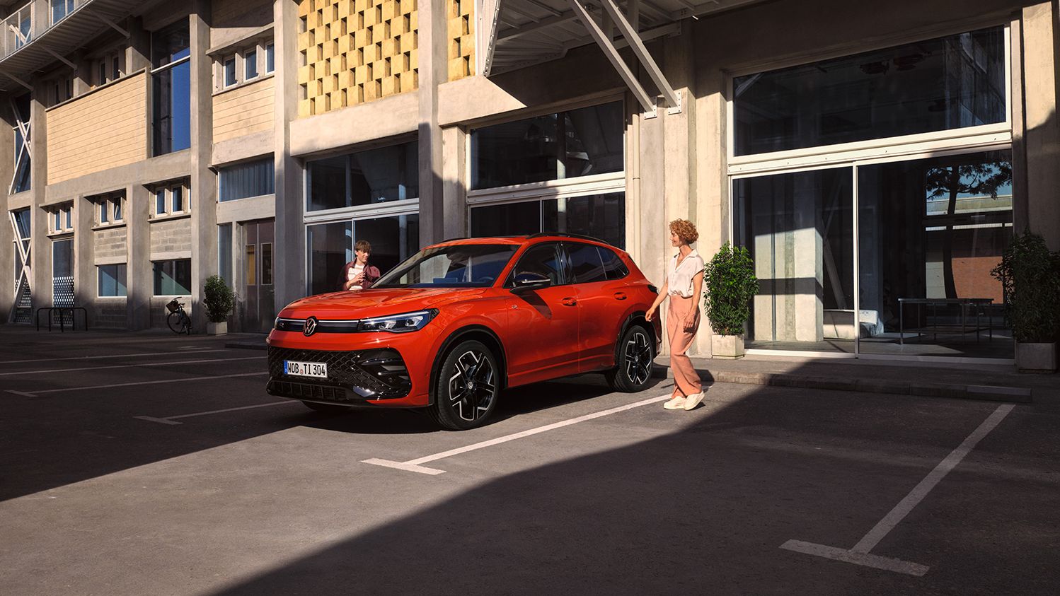 Vista lateral de um VW Tiguan vermelho, parado em frente a um edifício, uma jovem passa do lado do condutor.