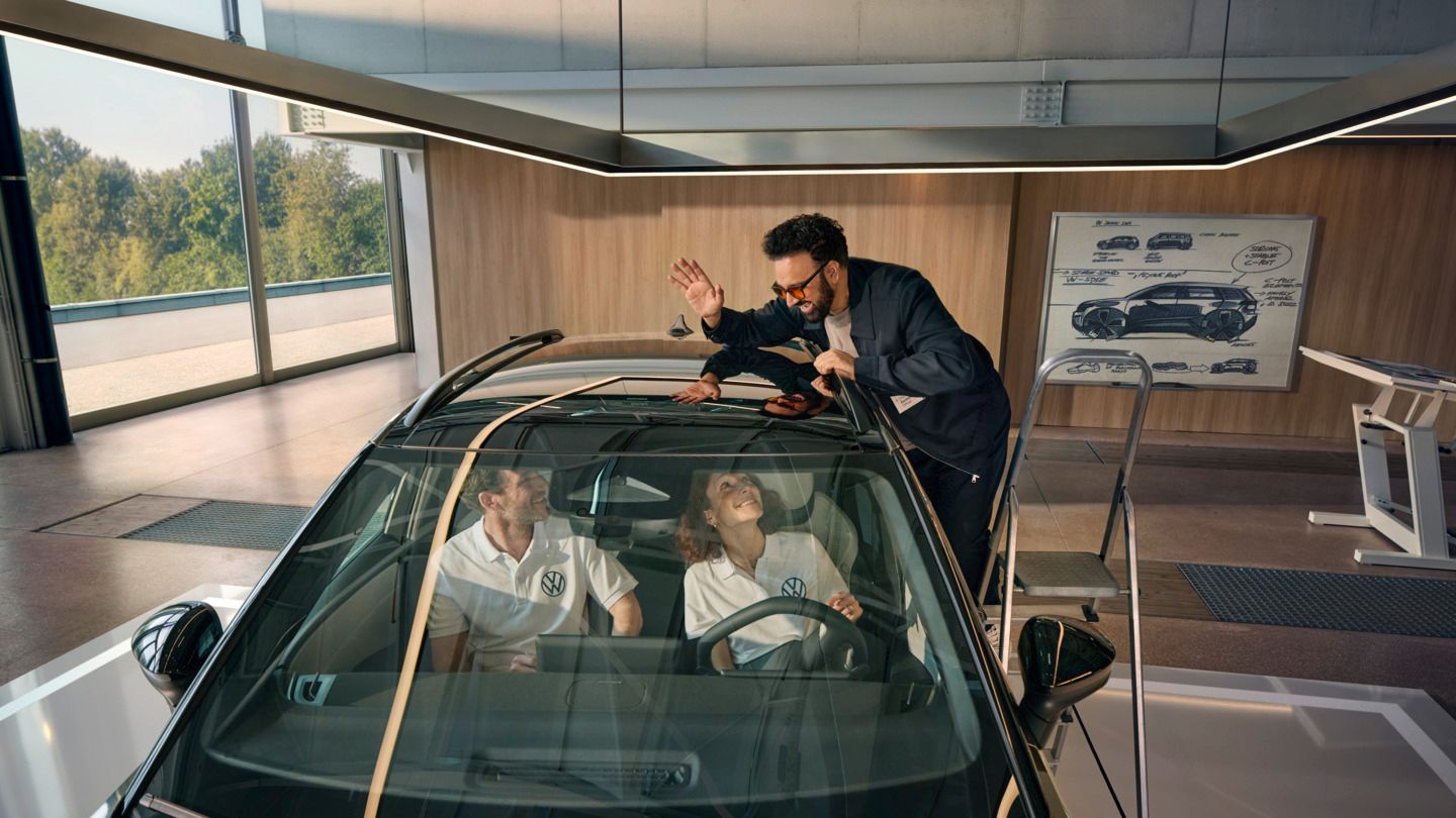 Um homem encontra-se numa escada ao lado de um VW ID. Cross a acenar pelo teto panorâmico às pessoas sentadas no seu interior.
