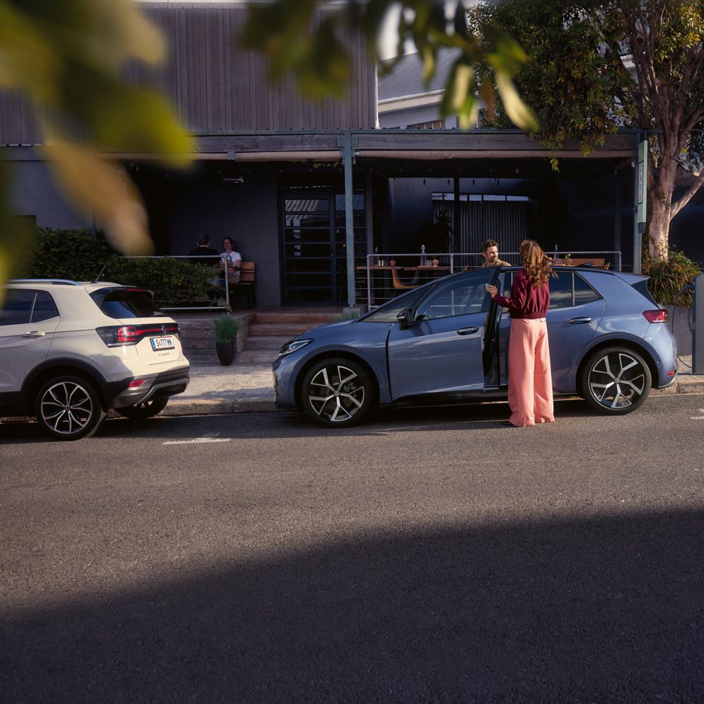 Eine Frau und ein Mann steigen in einen blauen VW ID.3 Neo, der vor einem Café parkt