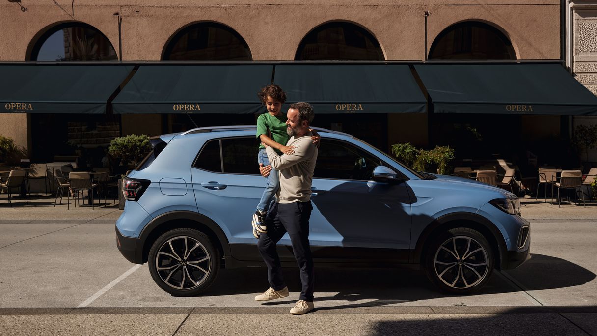 Um homem segura um menino alegre nos braços perante um VW T-Cross azul.