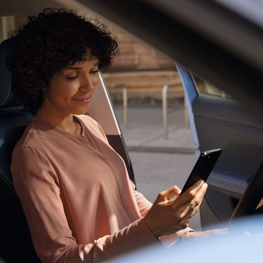 Uma mulher sorridente olha para o telemóvel no banco do condutor de um VW T-Cross estacionado.
