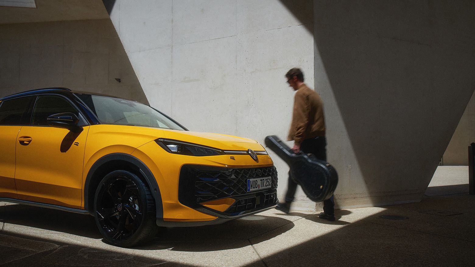Um homem com um estojo de guitarra passa por um VW T-Roc R-Line amarelo num ambiente de arquitetura moderna.