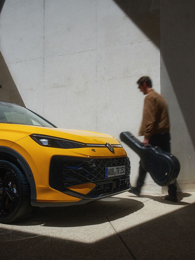 Um homem com um estojo de guitarra passa por um VW T-Roc R-Line amarelo num ambiente de arquitetura moderna.