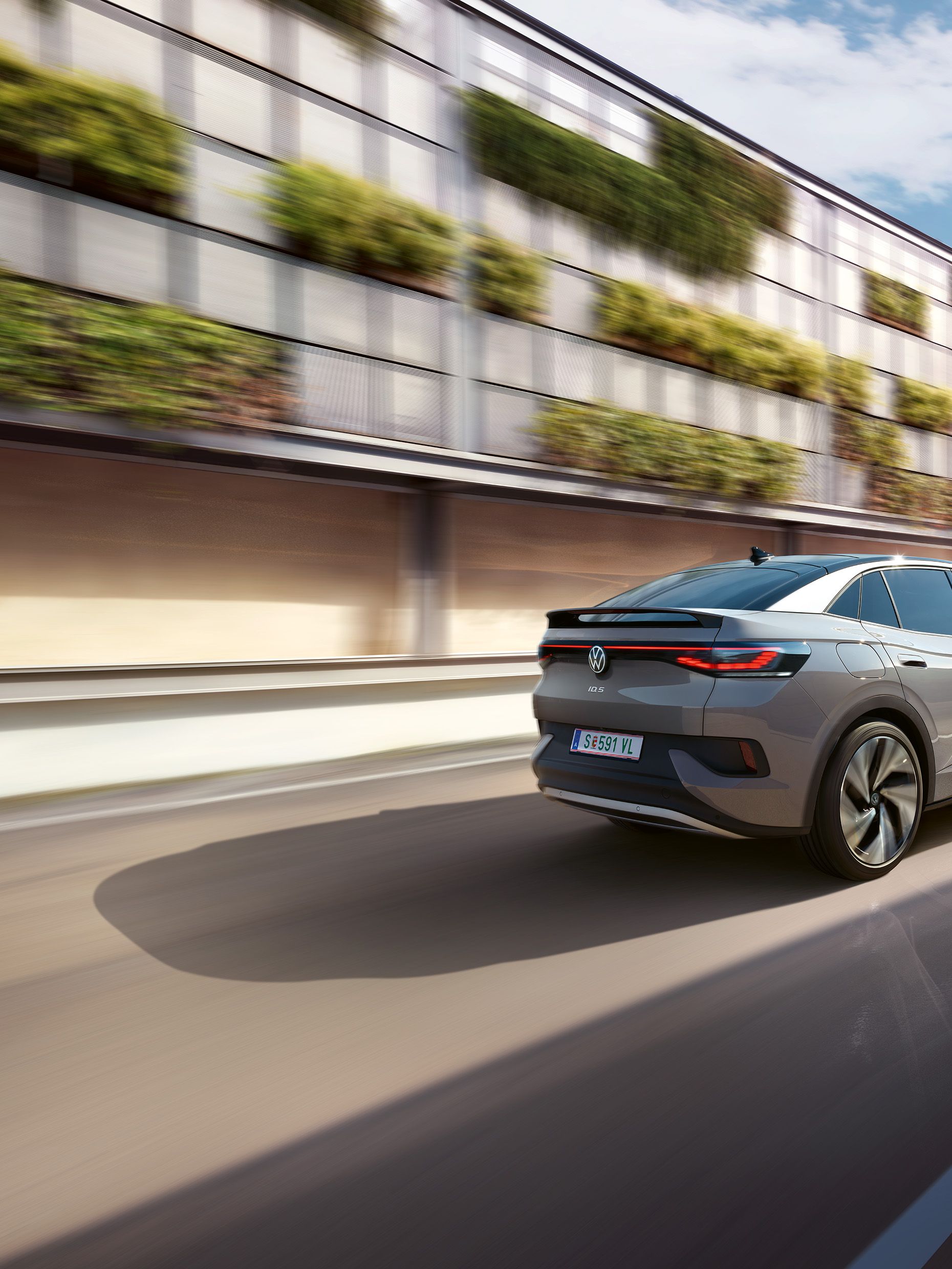 Fahraufnahme eines grauen VW ID.5 von schräg hinten, links ein helles, teilweise grün bewachsenes Gebäude