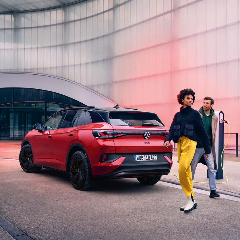 Um VW ID.4 GTX vermelho com o cabo de carregamento conectado em frente de um edifício cinzento com um homem e uma mulher a dirigir-se para o carro.