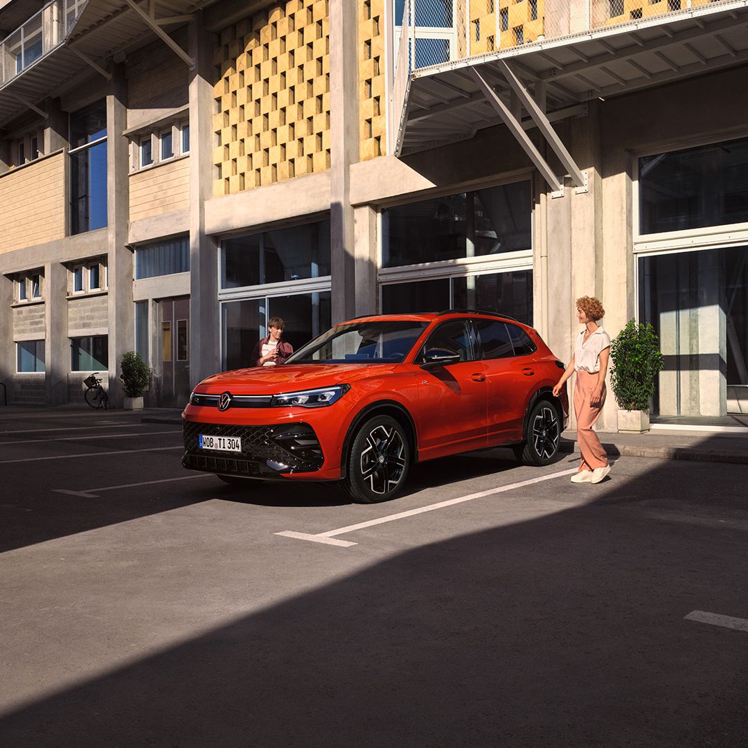 Vista lateral de um VW Tiguan vermelho, parado em frente a um edifício, uma jovem passa do lado do condutor.