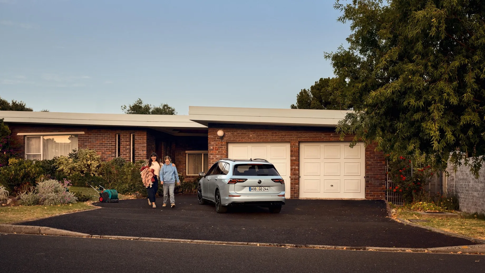 Weißer VW Golf Variant parkt vor der Garagentür eines Hauses. Eine Frau und ihr Kind gehen aus dem Haus raus.