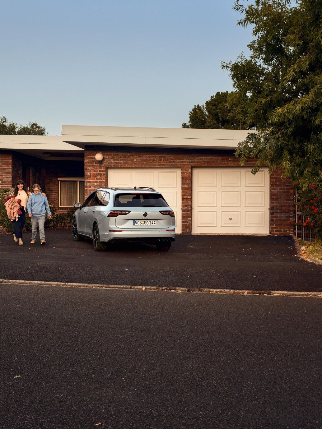 Weißer VW Golf Variant parkt vor der Garagentür eines Hauses. Eine Frau und ihr Kind gehen aus dem Haus raus.