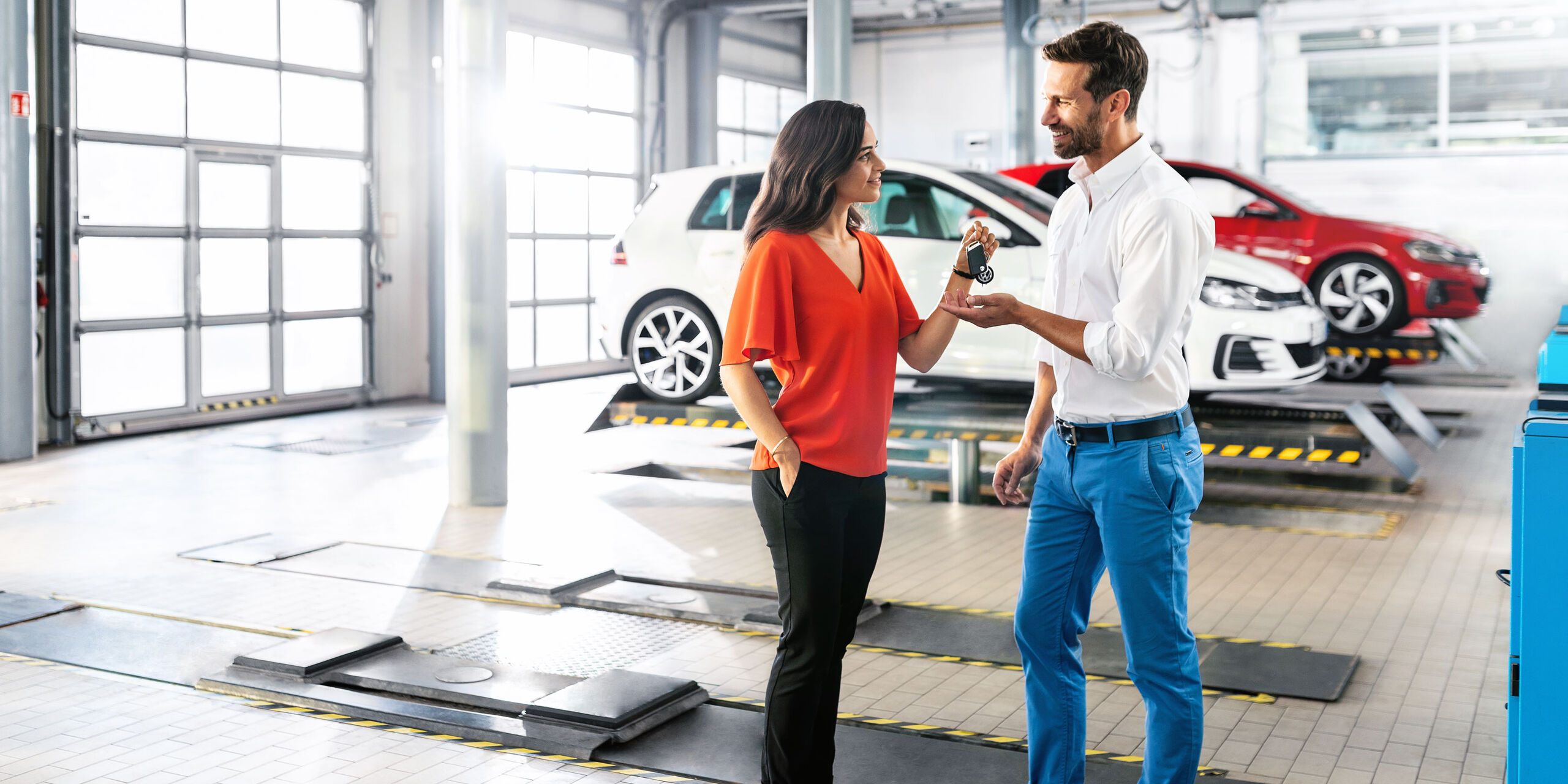 Eine Frau übergibt Ihren Autoschlüssel in einer Autowerkstatt an einen Mann. Im Hintergrund sind Volkswagen zu sehen.