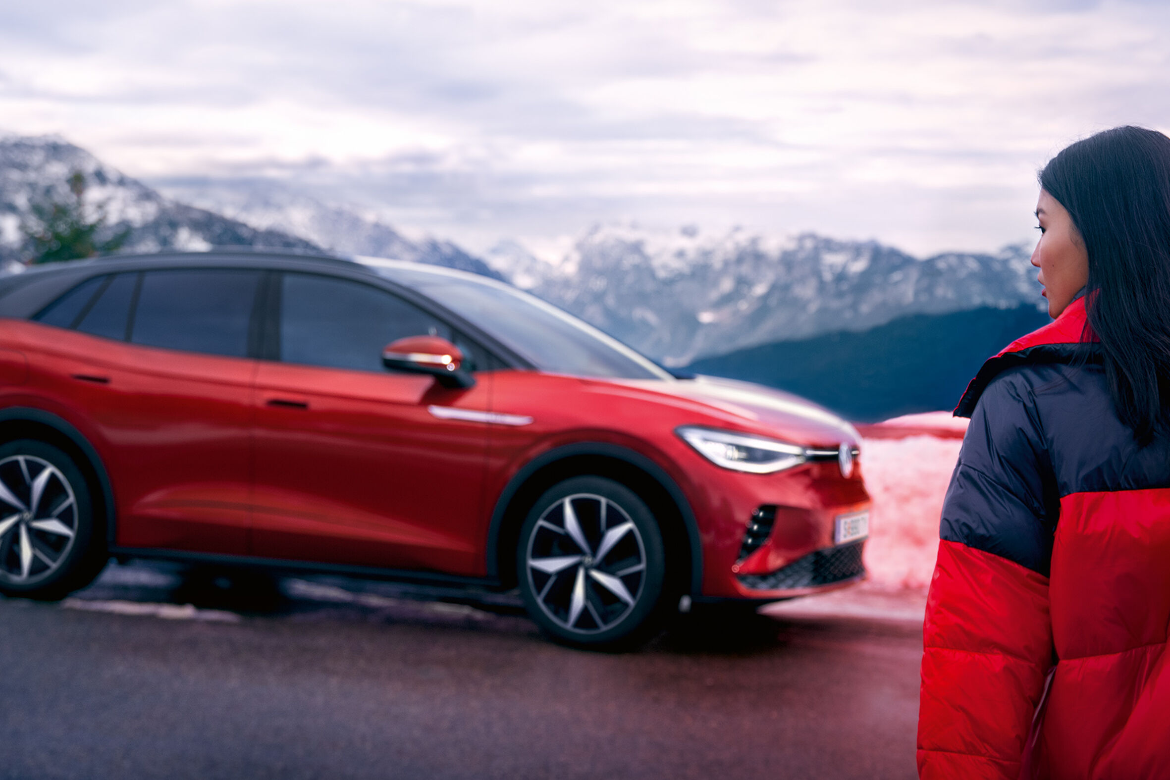 VW ID.4 GTX vermelho vísivel de lado. De pé na berma da estrada em frente às montanhas, está uma jovem mulher com uma prancha de snowboard a caminhar na sua direção.