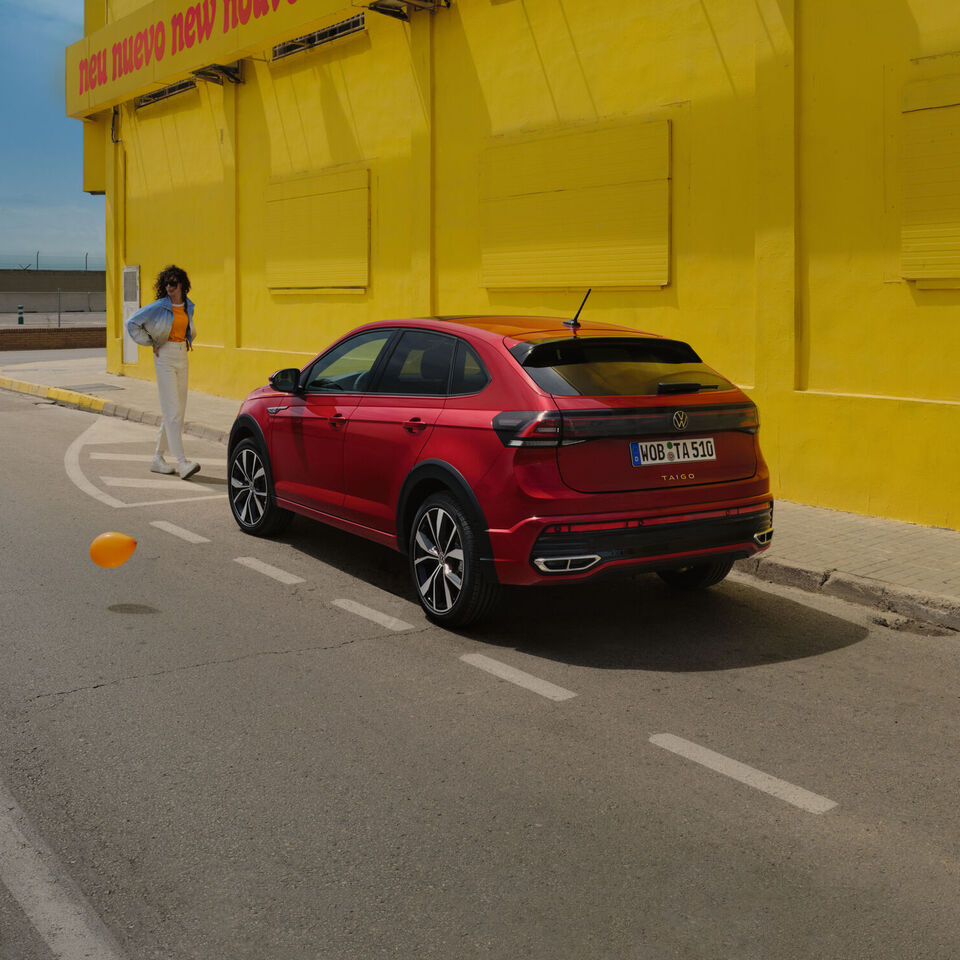 VW Taigo em vermelho na berma da estrada, em frente a um edifício amarelo, vista traseira e lateral, uma mulher dirige-se para o veículo