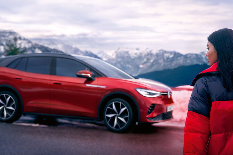 VW ID.4 GTX vermelho vísivel de lado. De pé na berma da estrada em frente às montanhas, está uma jovem mulher com uma prancha de snowboard a caminhar na sua direção.