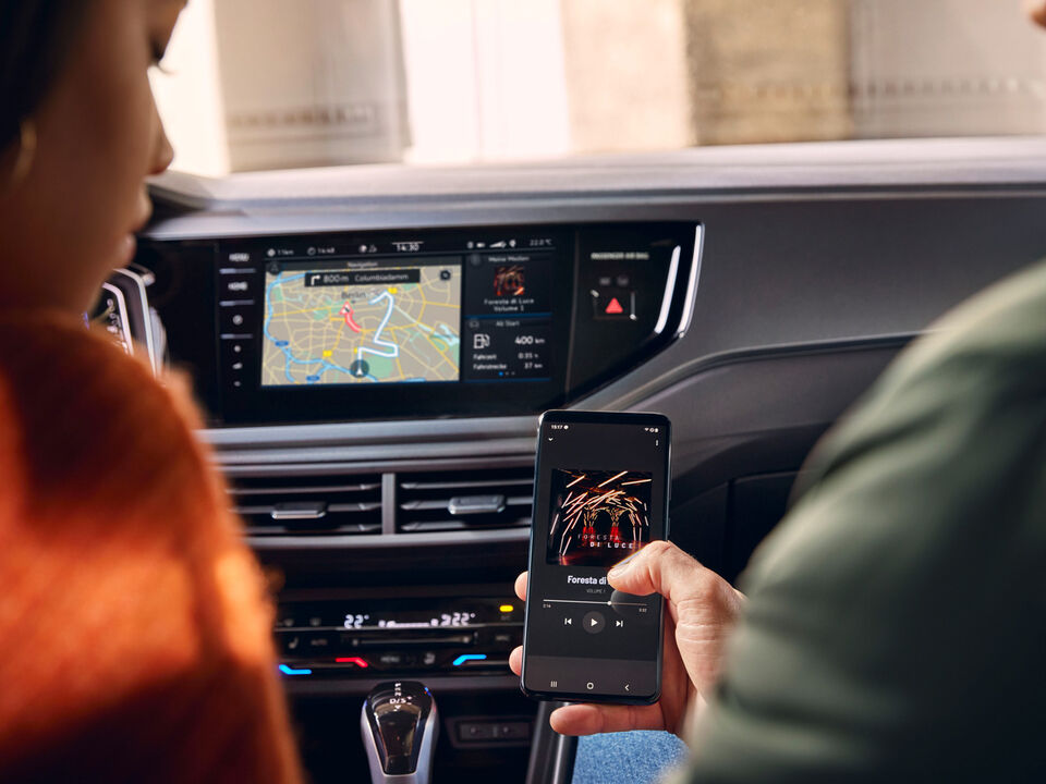 Duas pessoas sentadas à frente num VW Polo, no lado direito, uma delas segura um smartphone que se liga sem fios ao sistema de Infotainment. 