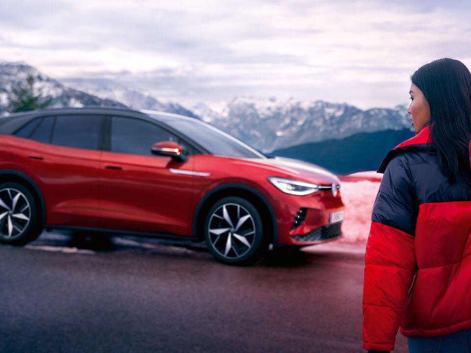 VW ID.4 GTX em vermelho na estrada numa paisagem de montanhas com neve. Mulher de casaco vermelho e preto atravessa a estrada com esquis e olha para a esquerda.