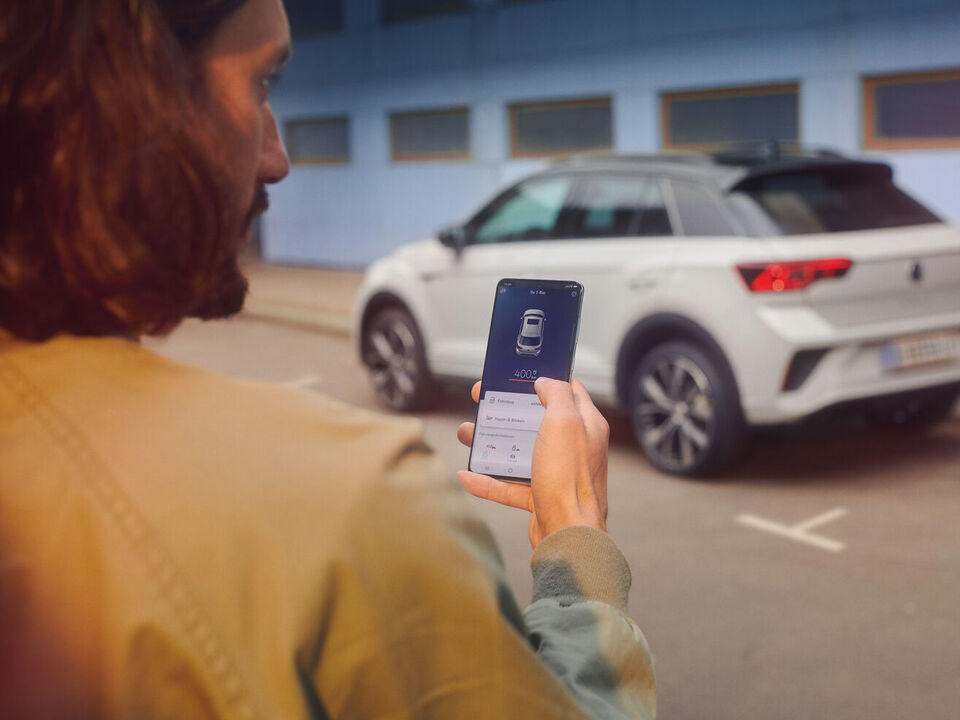 VW T-Roc R-Line em branco em frente de um edifício, vê-se a lateral, um homem em primeiro plano com smartphone na mão