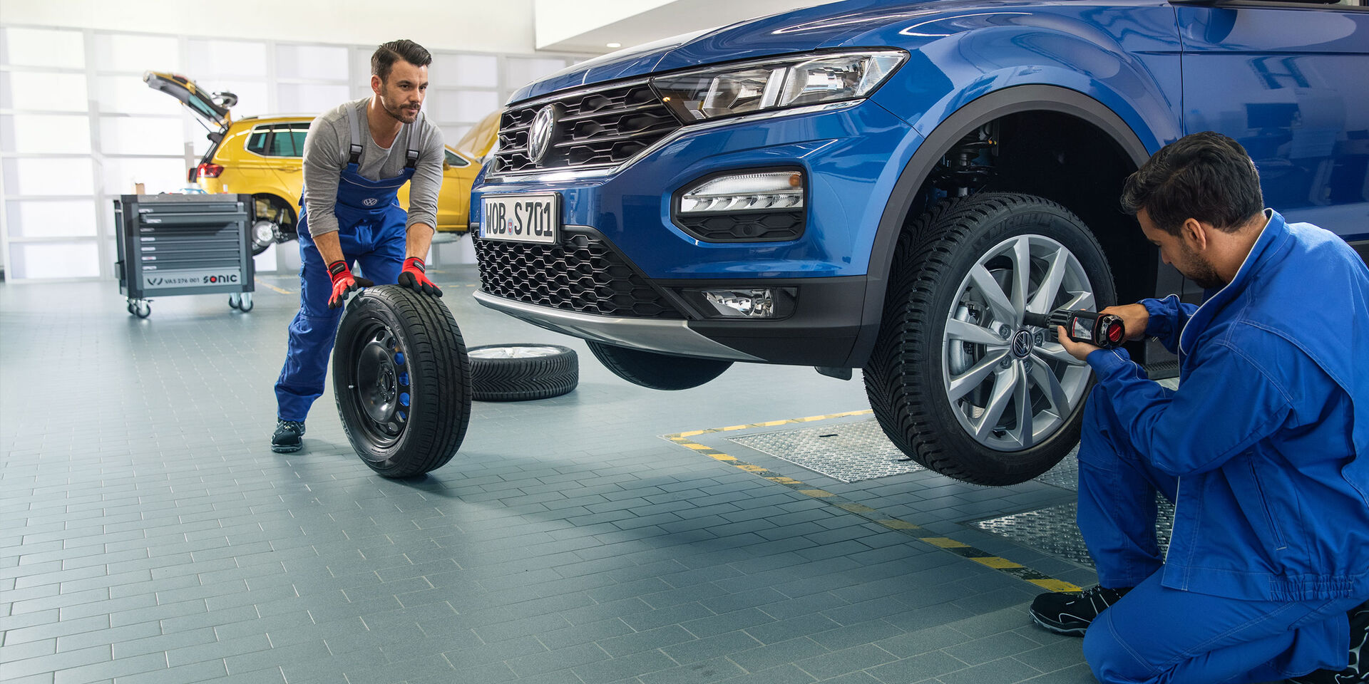 Dois empregados do serviço VW trabalham juntos na troca de pneus de um VW Tiguan azul numa oficina