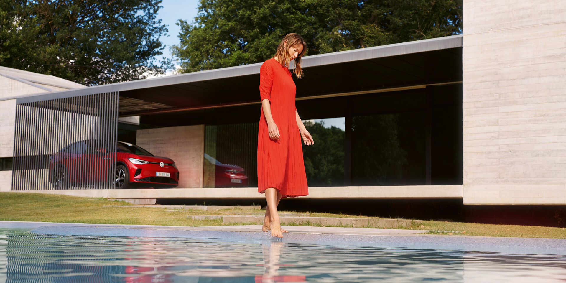 VW ID.5 GTX vermelho a estacionar em frente de uma casa. Mulher de vestido vermelho junto à piscina.