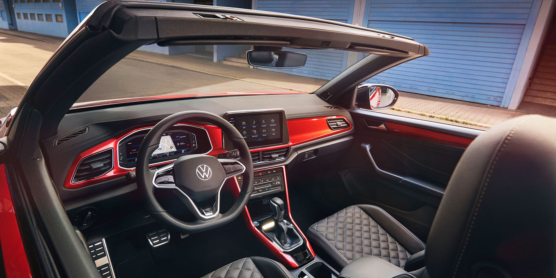Interieur des VW T-Roc Cabriolet: Blick durch das geöffnete Dach von oben auf das Cockpit mit rotem Dekor und die vorderen Sitze. 
